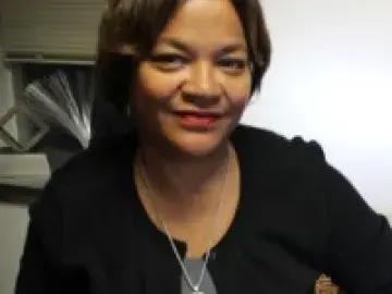 Nona Cheeks in black top and necklaces, seated indoors near blinds