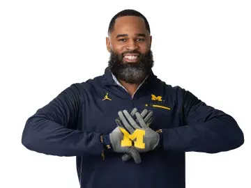 Jason Harris in navy sweatshirt and gloves forming an “M” hand sign, studio portrait
