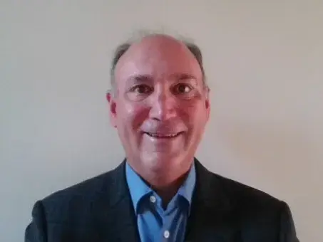 Jim Galante smiling in blazer and blue shirt against plain wall.
