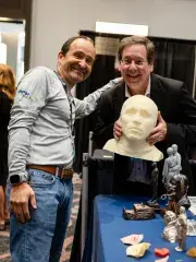 Two men smile at conference booth with mannequin head and tech devices on table.